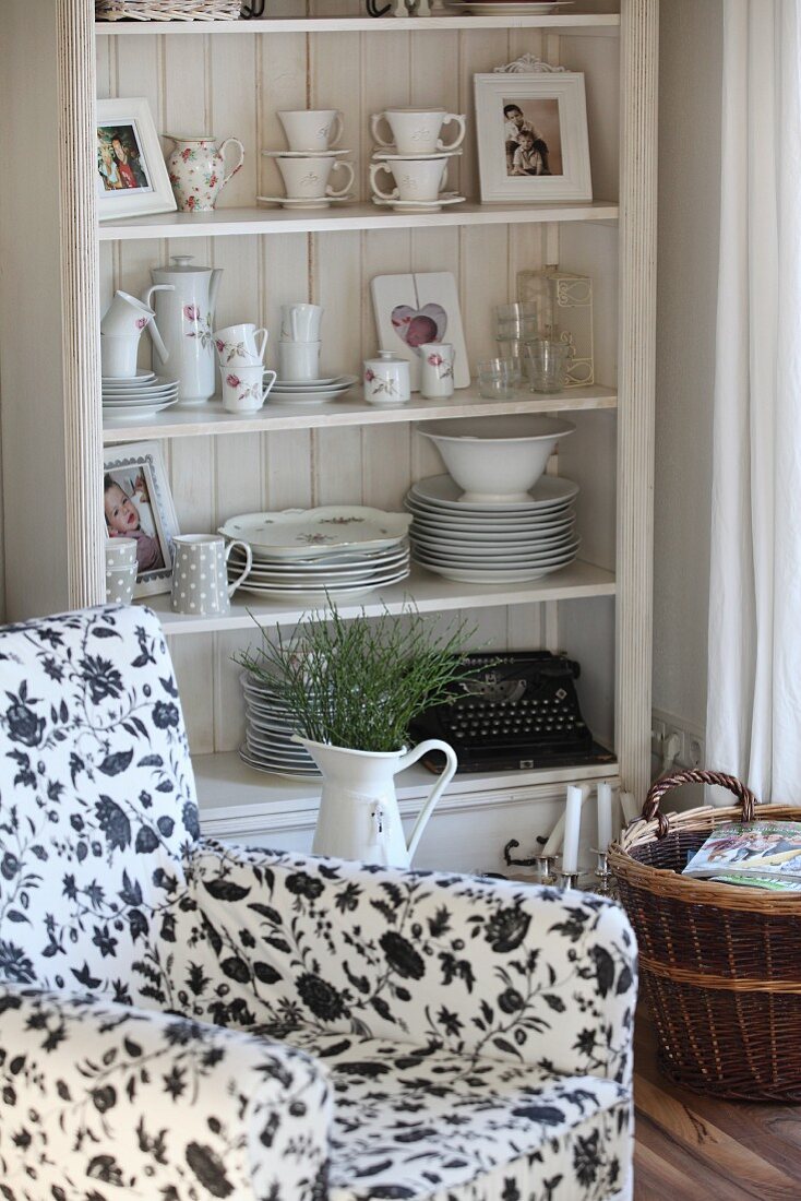 Crockery on country-house shelves behind enamel jug and armchair with black and white floral upholstery