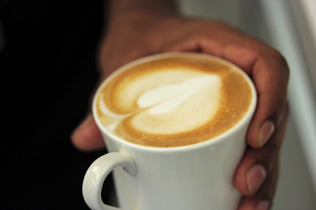 A hand holding a cup of cappuccino with a heart-shaped pattern in the foam