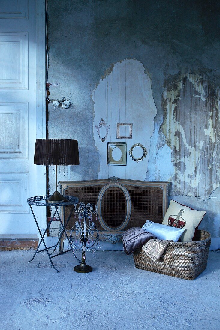 Table lamp with black lampshade on side table next to candelabra and basket on floor in front of wall with crumbling plaster