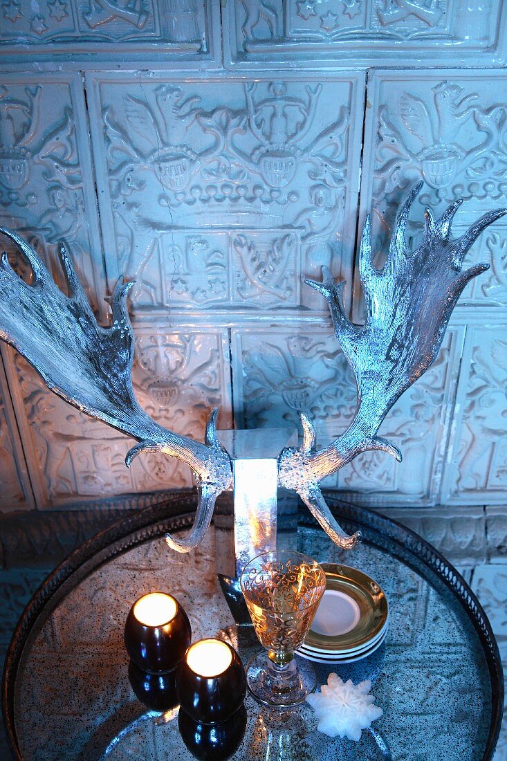 Tealights and silvered stag's antlers on tray in front of tiled wall with antique embossed motifs