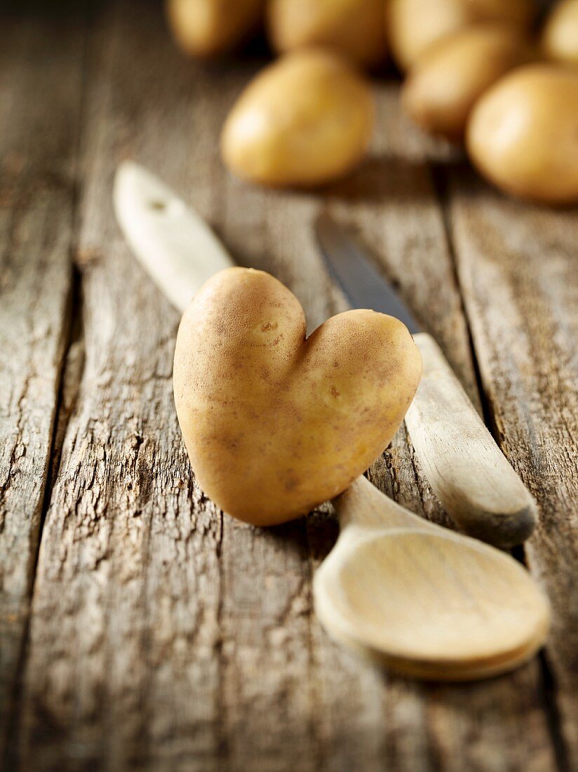 Herzförmige Kartoffel, Kochlöffel und Messer auf Holzuntergrund