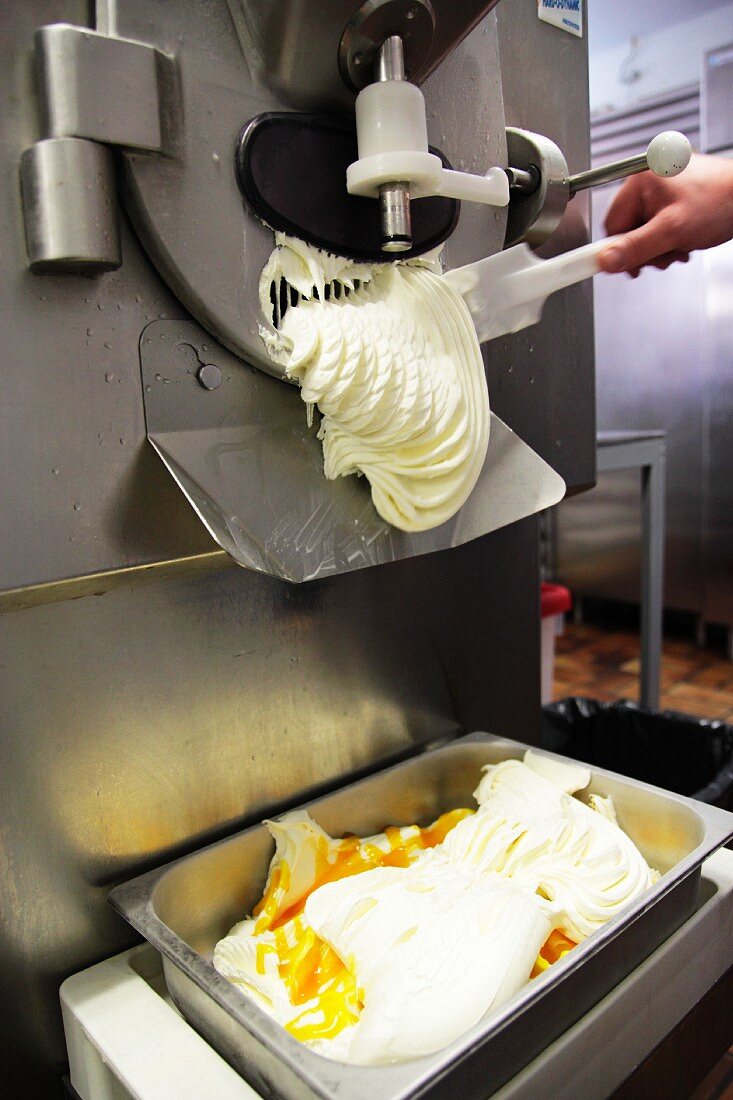 An ice cream machine producing a pale ice-cream mixture, a hand helping to release the ice cream with a spatula