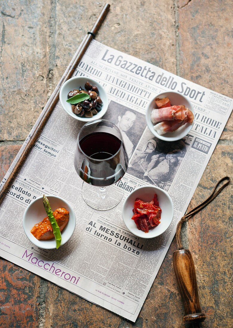 Assorted antipasti and a glass of red wine on a newspaper (Italy)