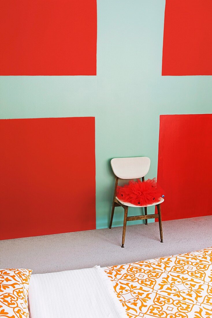 Red fabric on chair against wall painted with graphic design in red and turquoise beyond bed with ornamental patterned bed linen