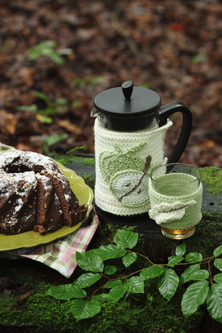 Crocheted coffee pot and glass warmer next to chocolate cake on plate in woodland