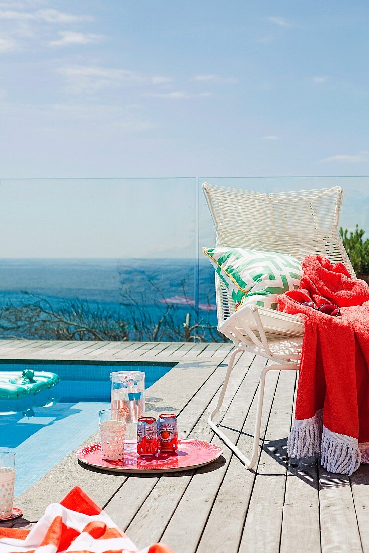 Erfrischungsgetränke auf Holz-Terrassenboden neben weißem Flechtstuhl am Pool, im Hintergrund Glasbrüstung und Blick aufs Meer