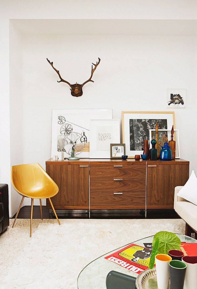 Schalenstuhl aus Schichtholz im Fifties Stil vor einem 70er Jahre Sideboard mit aufgestellten Bilder und farbigen Flaschen, darüber ein Hirschgeweih