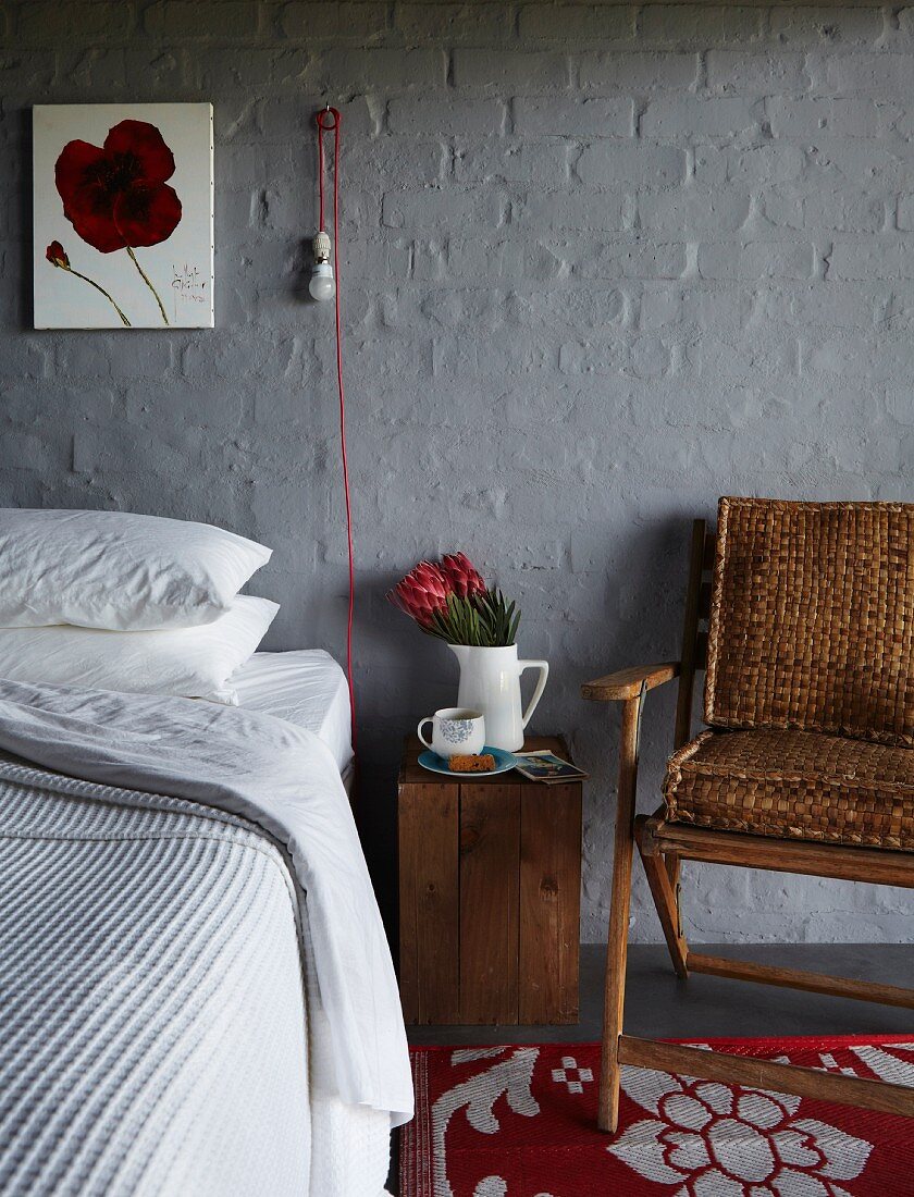 Bett an grau gestrichener Ziegelwand mit naturbelassener Holzkiste als Nachtkästchen, Armlehnstuhl und innovativer Wandleuchte