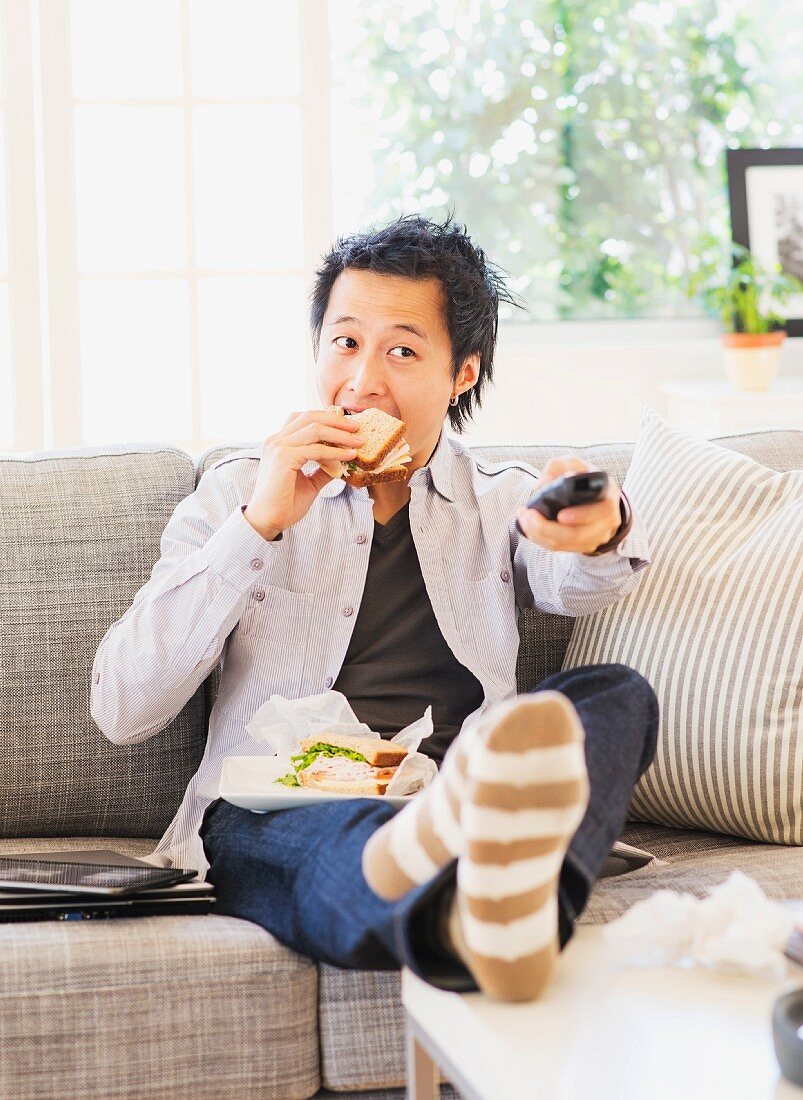 An Asian man sitting on the sofa in front of the television holding a sandwich and a remote control