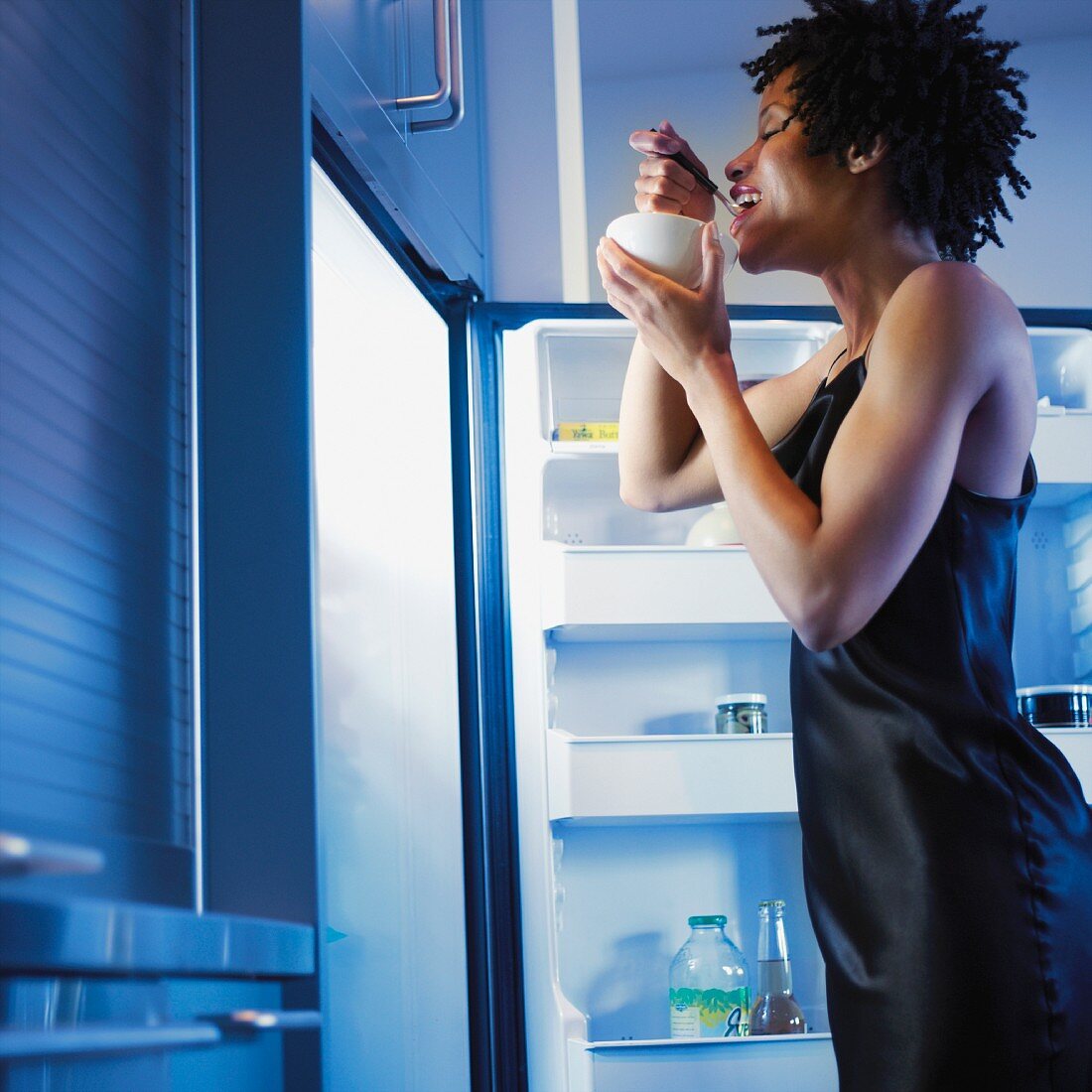 Afrikanische Frau isst neben dem offenen Kühlschrank