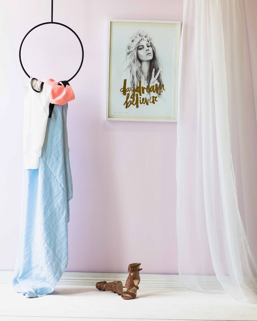 Ladies' sandals on white floor next to clothing hanging from circular metal frame suspended from ceiling, framed portrait of woman on wall and airy curtain to one side