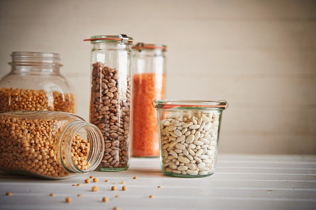 Assorted jars of beans and grains on wooden surface including corn, lentils, pinto beans, canellini beans, and peas