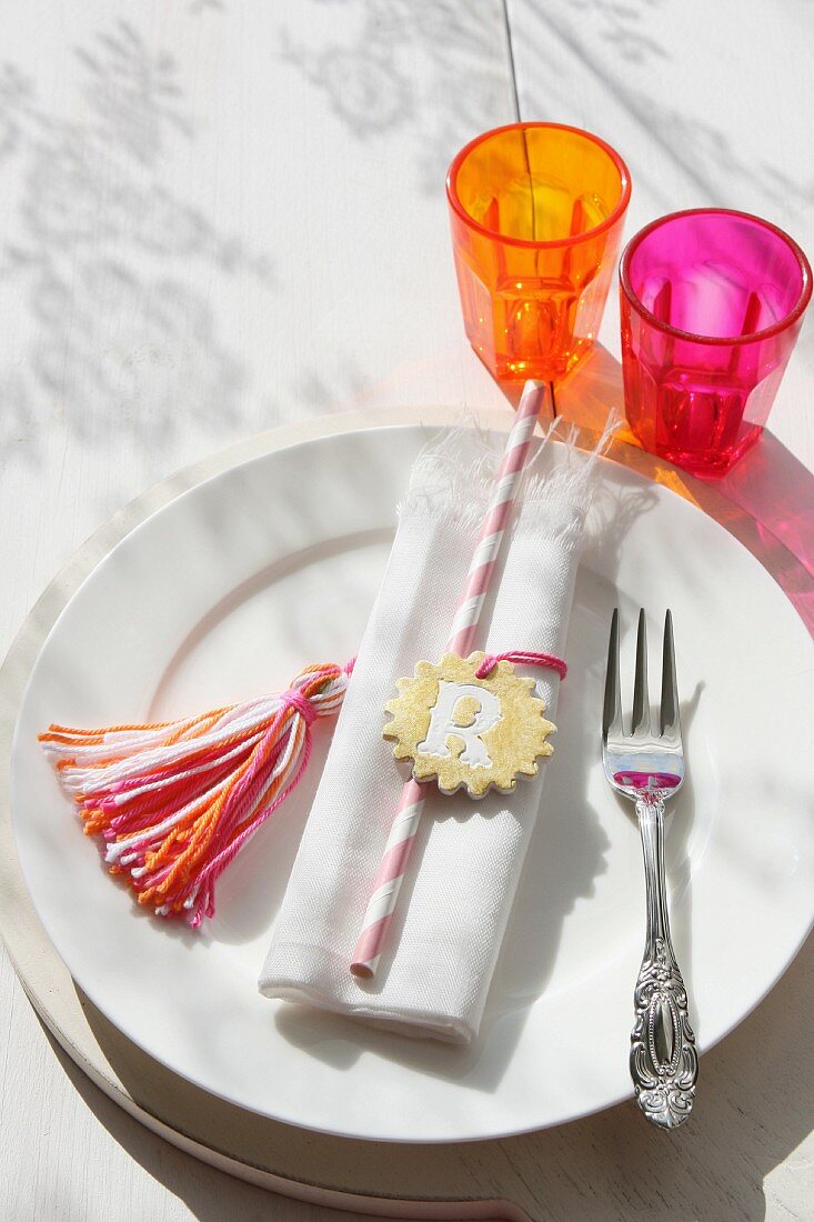 Napkin and clay pendant on white plate next to colourful glasses