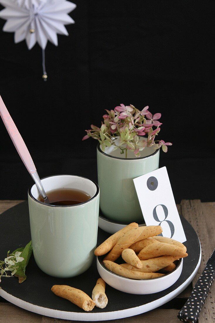 Knabbergebäck, Teetasse und Blütenstengel auf schwarzer Holzplatte vor dunklem Hintergrund