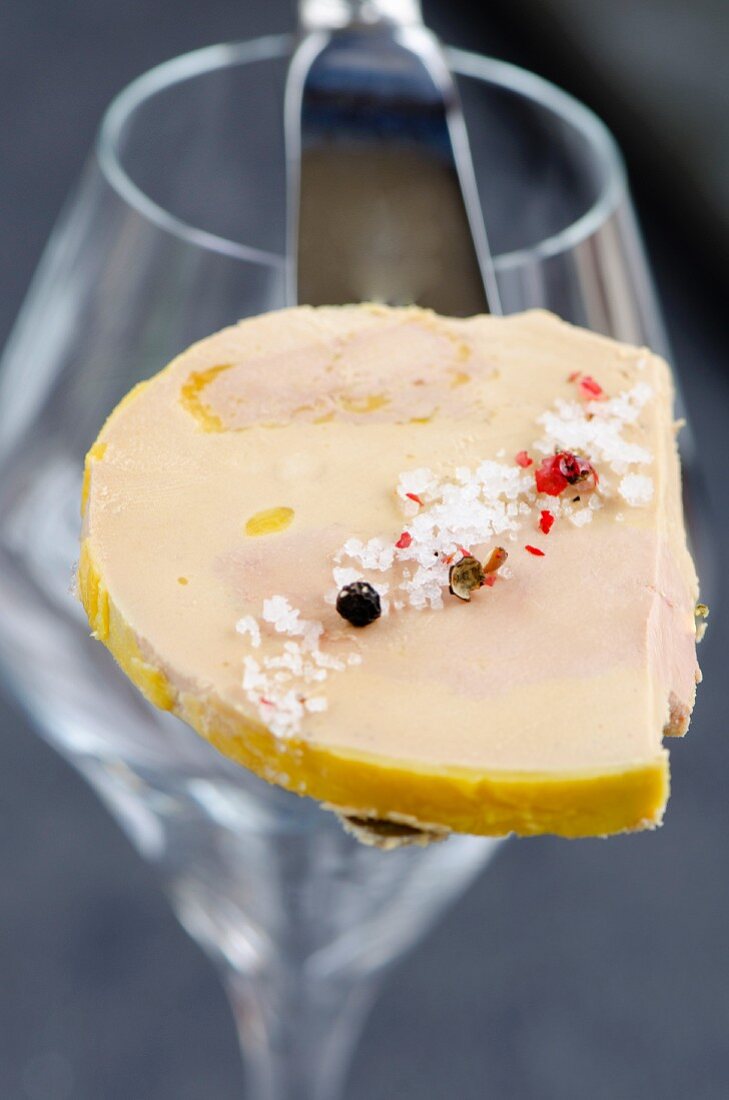 A slice of foie gras with fleur de sel and peppercorns, balanced on a knife, in front of a wine glass