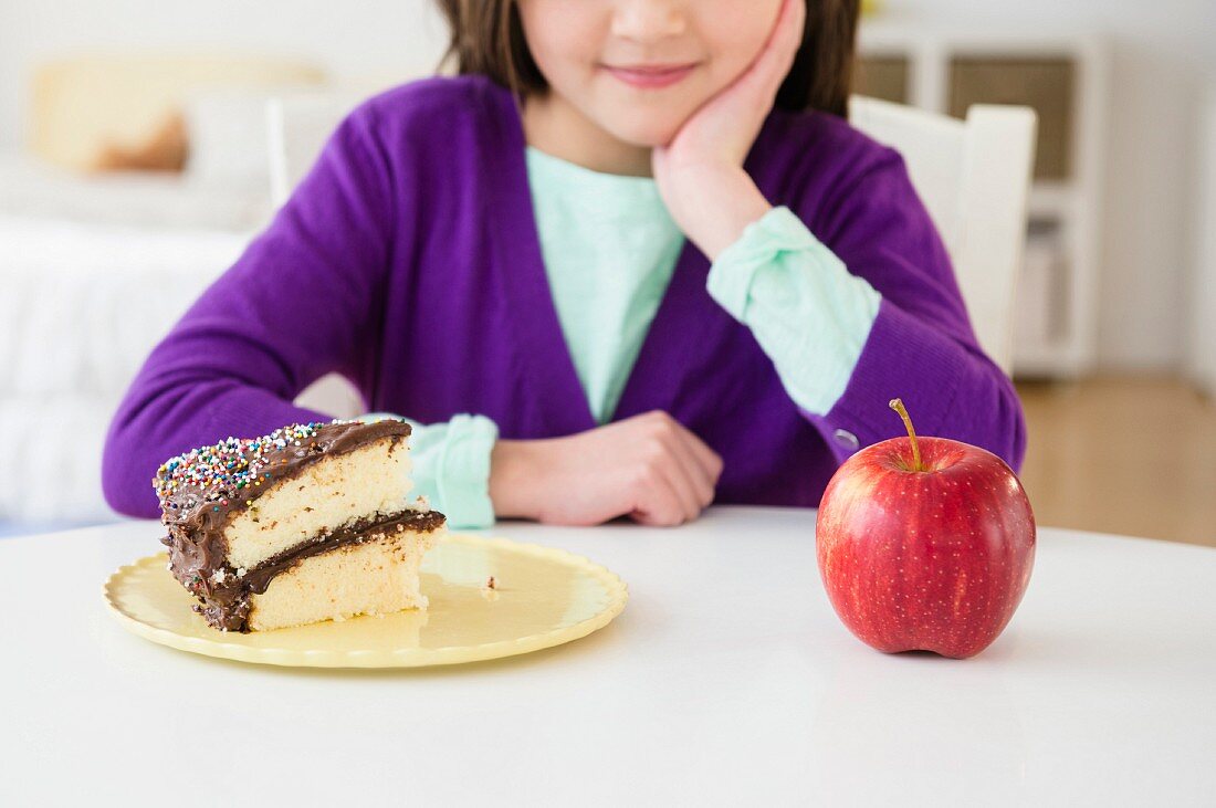 A girl sitting at a table in front of a slice of cake and an apple