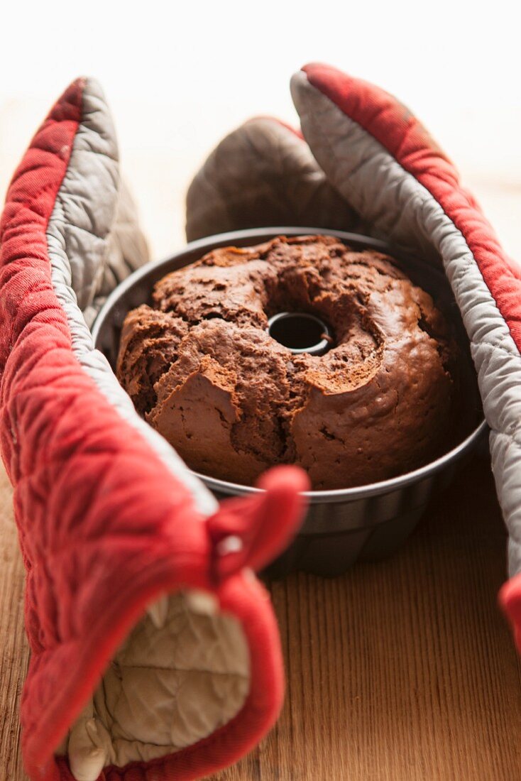 A Bundt cake in a baking tin between two oven gloves