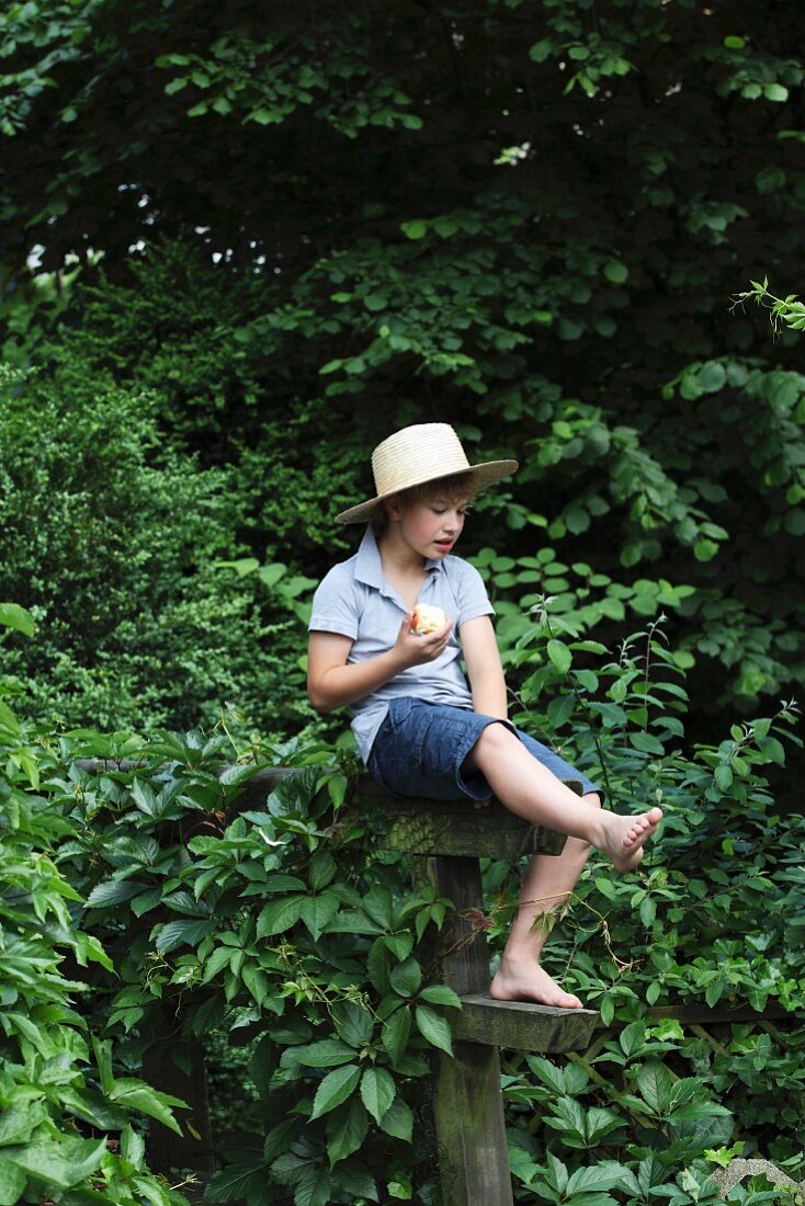 Boy outdoors wearing straw hat and summery clothing sitting on wooden structure