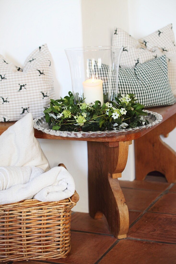 Lit candle in lantern and wintery wreath of mistletoe on tray between cushions on rustic stove bench