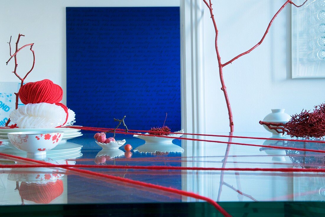 Asiatische Tischdeko: Roter Wollfaden um Glastisch gewickelt, dazwischen Schälchen und Tellerstapel mit Glasnudeln und roter Wollknäuel