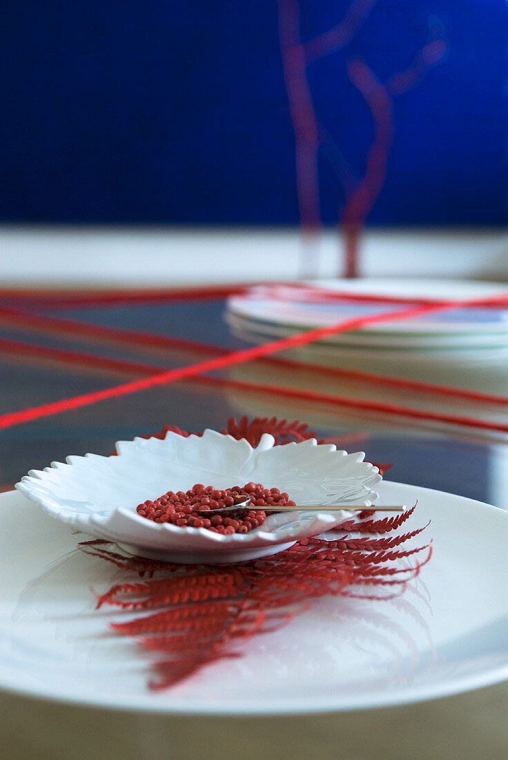 Asiatische Tischdeko: Blattförmiges Schälchen mit rotem Pfeffer auf rotgefäbtem Farnblatt und weißem Teller, roter Wollfaden umwickelt Tisch