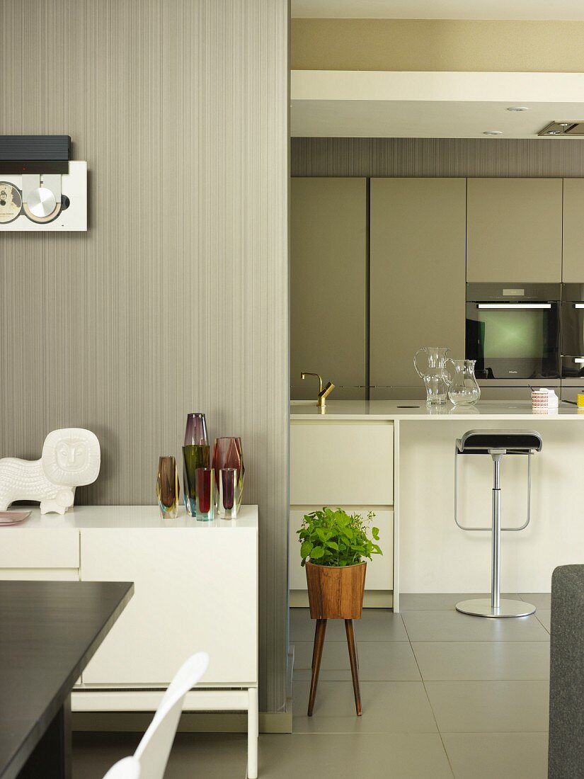 Detail of dining area with white sideboard against wooden wall varnished pale grey; modern, white fitted kitchen with bar stools at counter in background