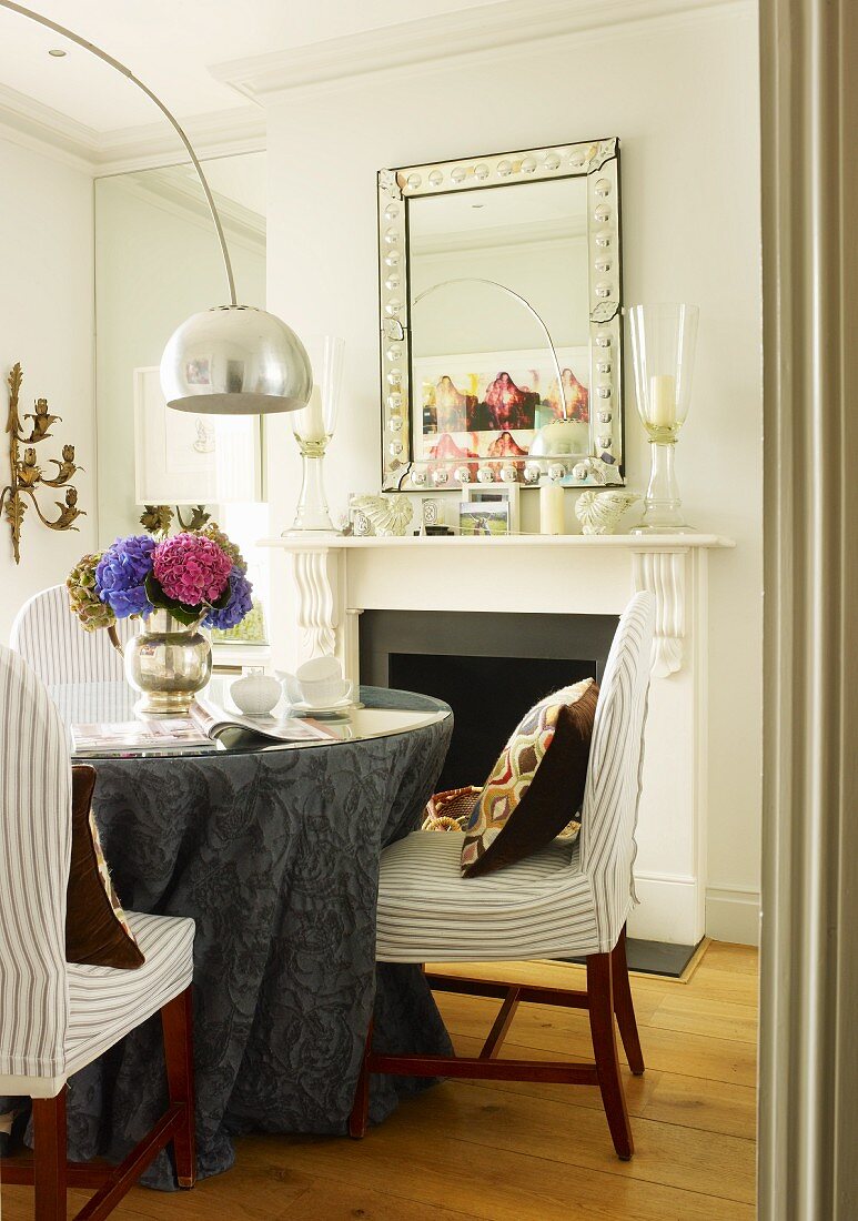 Dining area with grey damask tablecloth on round table and designer arc lamp; vintage mirror above open fireplace in background