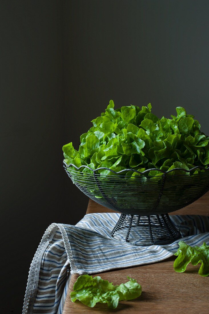 Frischer Salat im Drahtkorb auf Holztisch mit Geschirrtuch