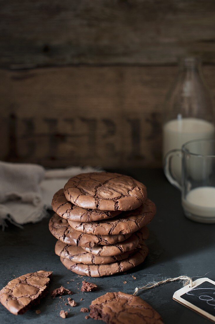 a stack of dark chocolate cookies one crumbled and a bottle of milk and a cup in a background.
