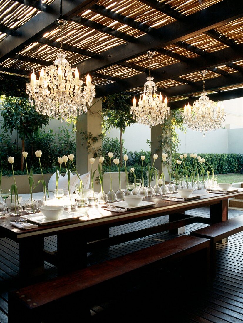 Outdoor table with integrated benches on roofed wooden terrace; table festively set with row of white tulips below chandeliers