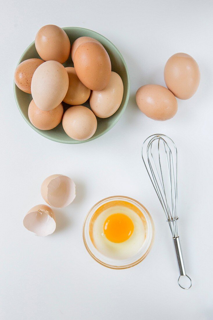 Frische Eier in einer Schüssel, aufgeschlagenes Ei und Schneebesen