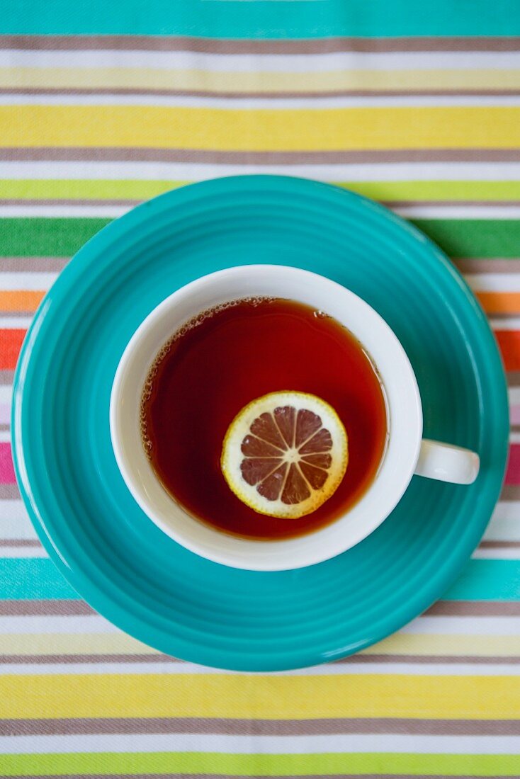 Teetasse mit Zitronenscheibe auf blauem Teller und gestreiftem Tischtuch