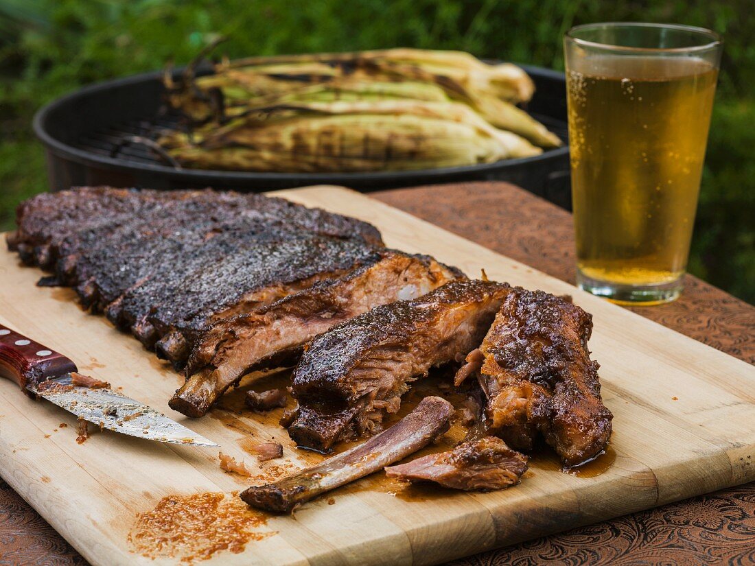 BBQ ribs outside on a cutting board with the first few segments cut exposing the meat with gilled corn and a beverage in the background