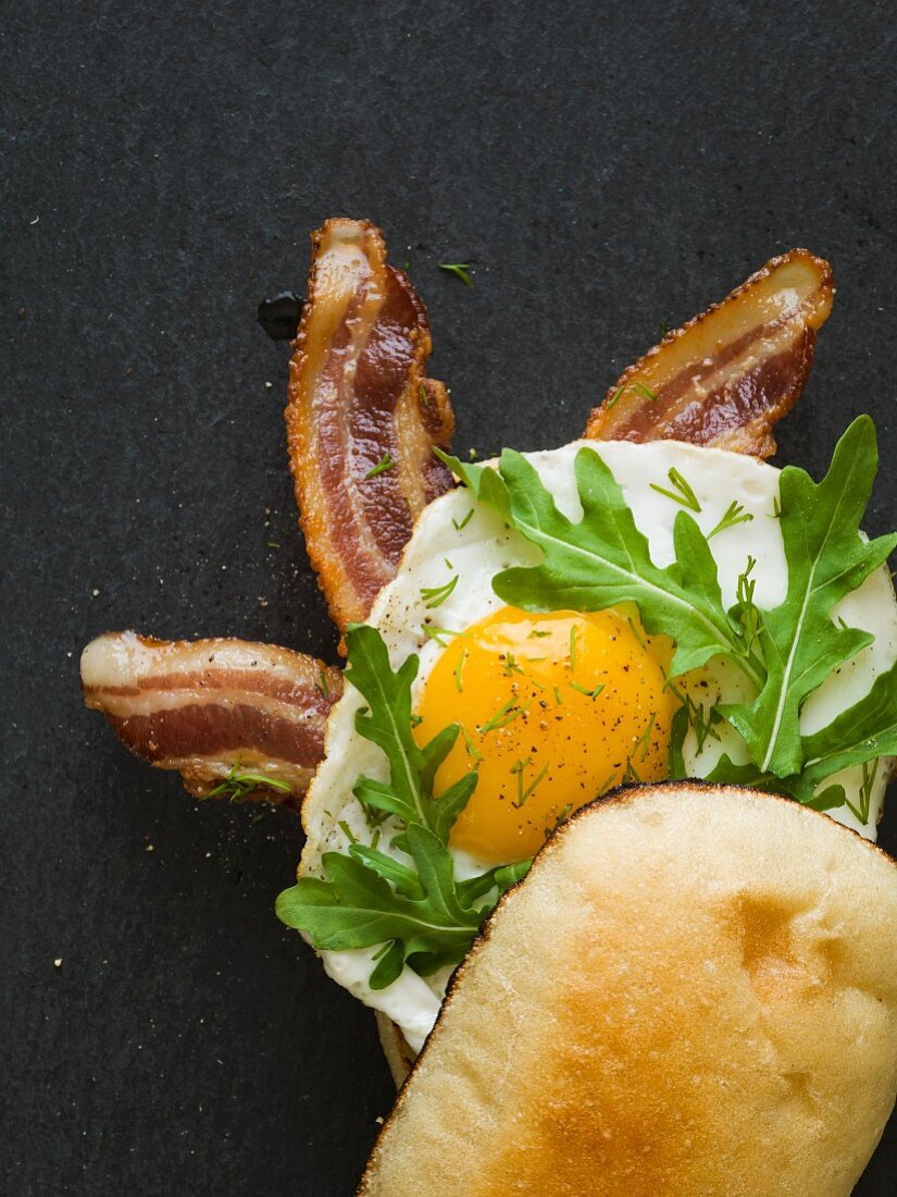 Breakfast sandwich with bacon, egg and arugula exposed sitting on a black slate cutting board