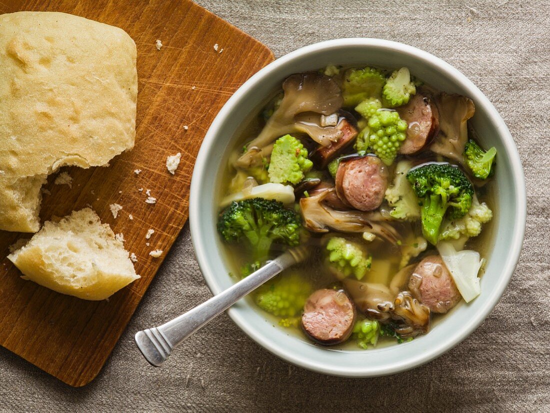 Overhead of soup with sausage, oyster mushrooms, broccoli and romanesco broccoli in a light blue bowl with bread on a small cutting board
