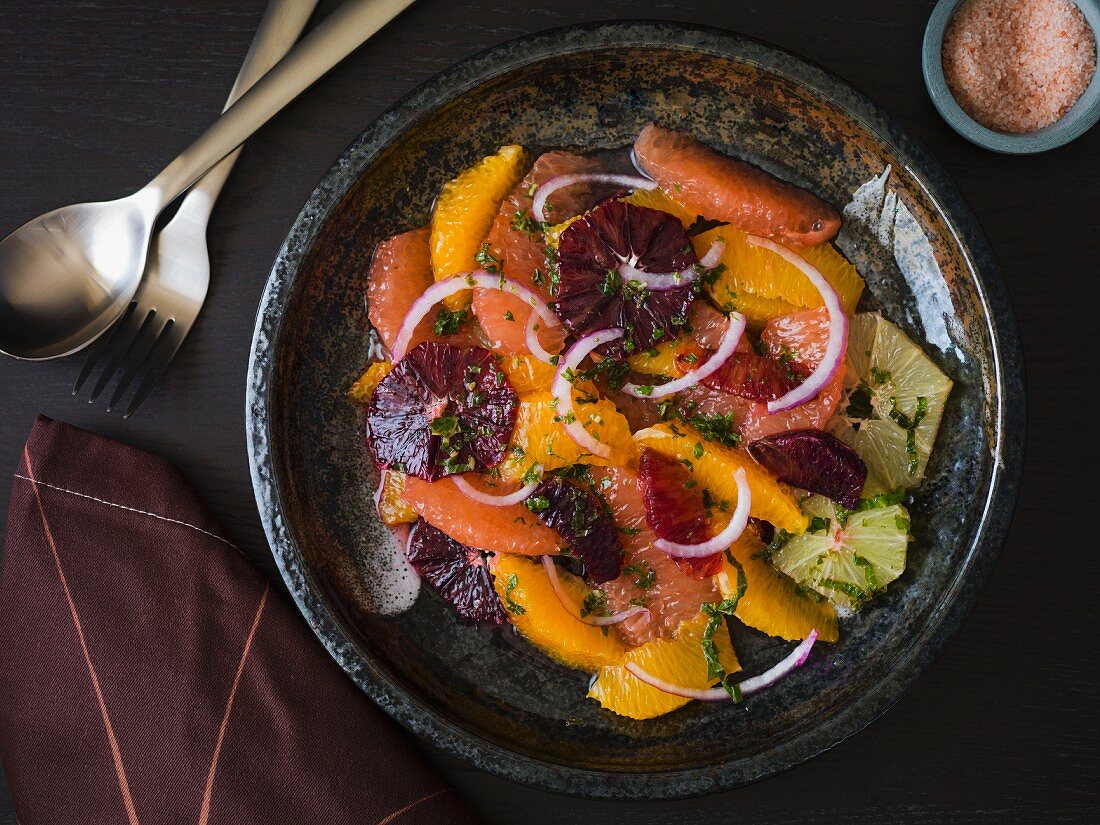 Citrus salad with grapefruit, orange, lime, mint and red onions in a dark stoneware plate with silver utensils on a dark surface