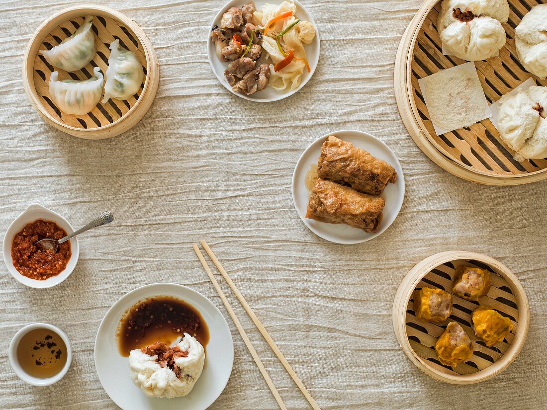 Overhead of bamboo steamers with various dim sum dumplings and sauces on a light material with chopsticks
