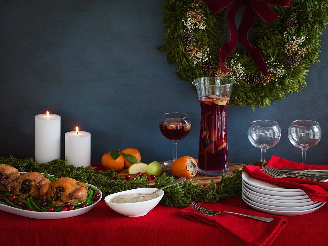 Holiday meal ready for serving with cornish game hens on a bed of green beans and cranberries, gravy and red sangria with stacked plates on a red tablecloth with white candles and a wreath in the background