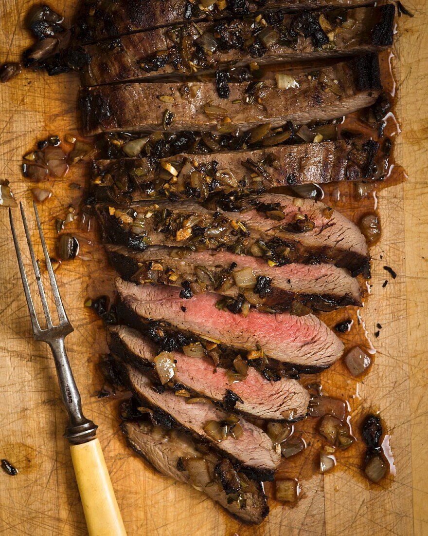 Overhead of London Broil steak sliced on a cutting board with carmelized onions and a vintage carving fork