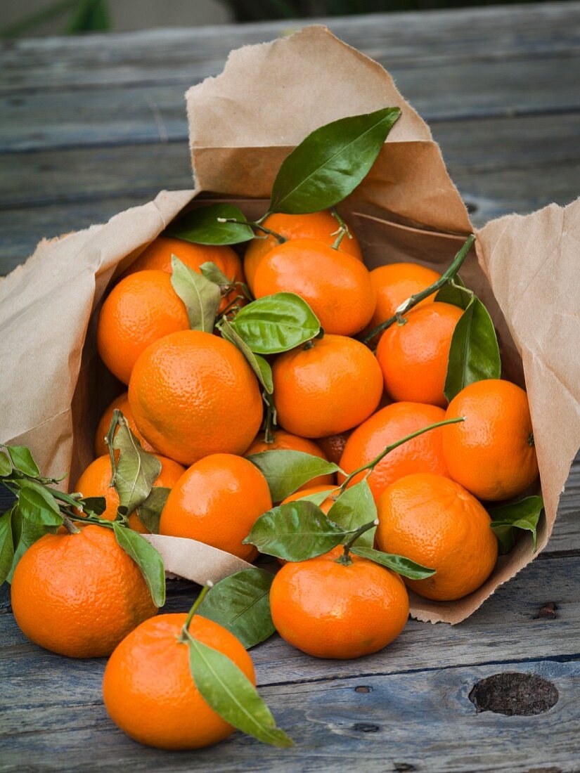 Clementine oranges with stems and leaves attached spilling out of a brown paper bag onto a rustic table