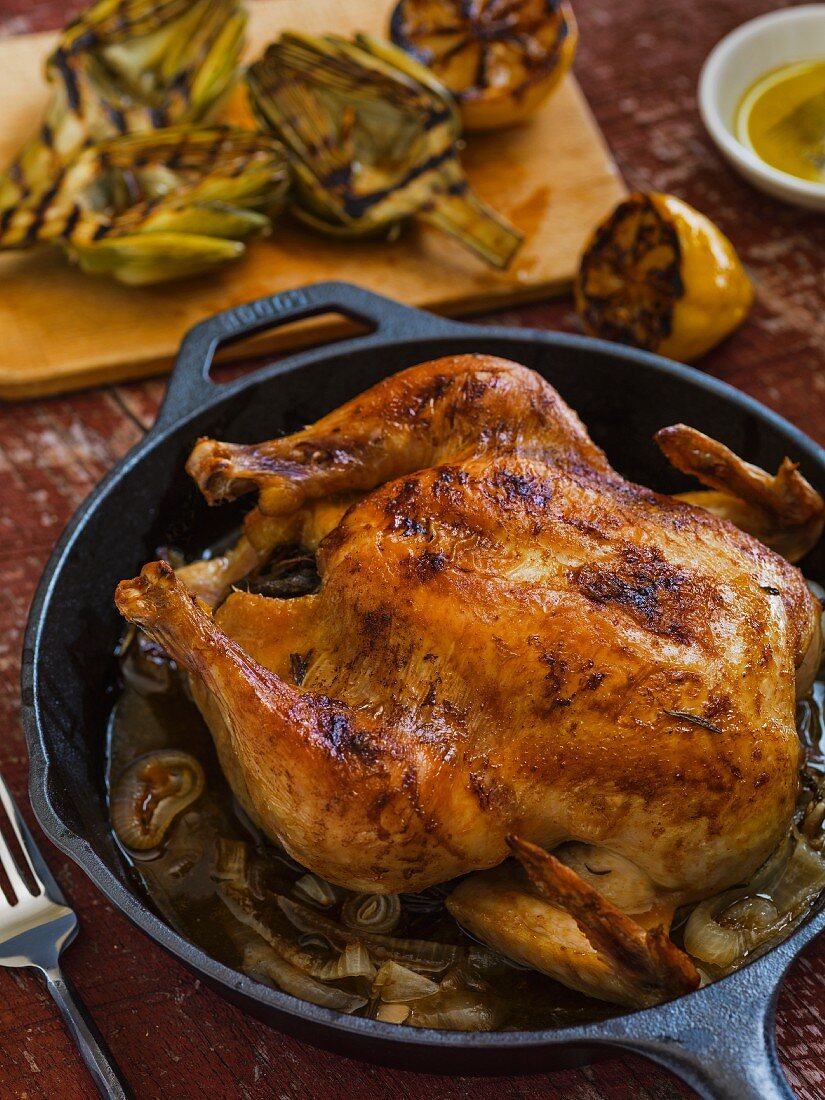 Whole roasted chicken in a cast iron pan on a rustic surface with grilled baby artichokes and lemon in the background
