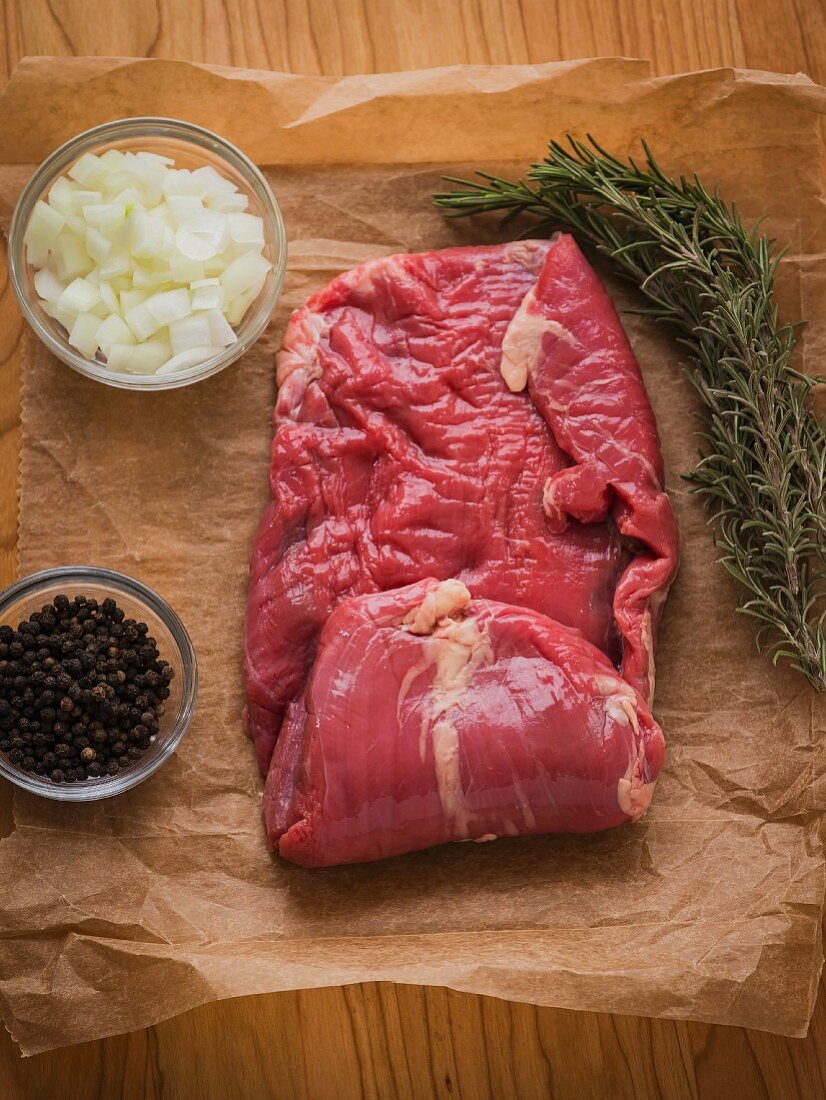 Raw skirt steak on brown butcher paper with rosemary sprigs, raw onions and black peppercorns