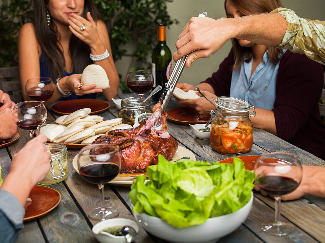Freunde geniessen Schweinebraten mit Brötchen, Salat, Kimchi und Rotwein an einem Gartentisch