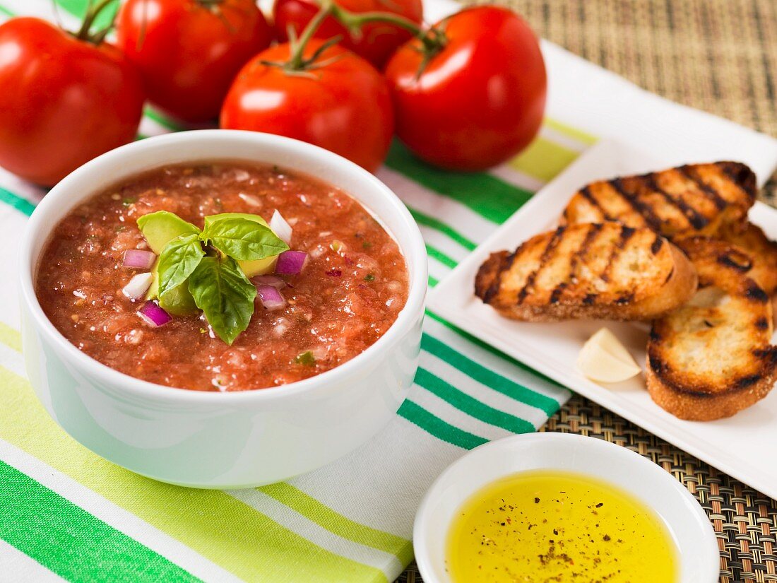 Gazpacho, garniert mit Avocado, roten Zwiebeln und Basilikum, daneben Olivenöl, Crostini und frische Tomaten