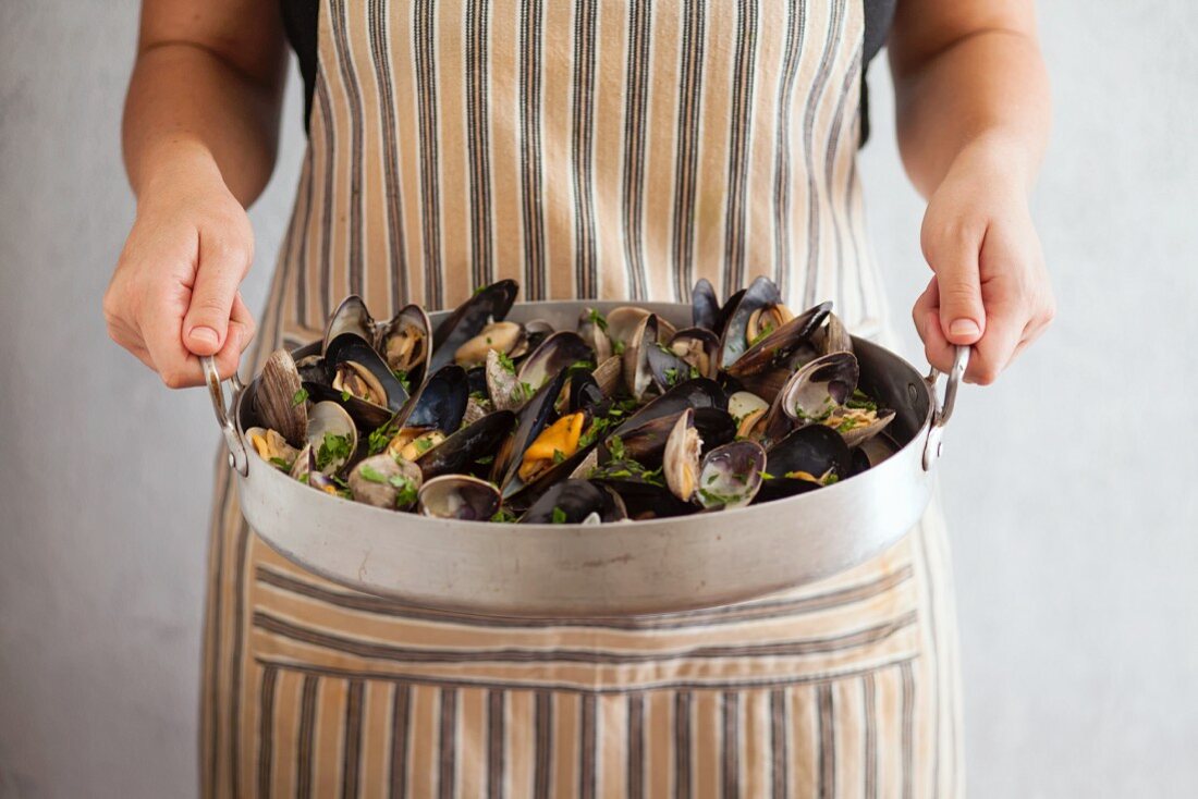 A person wearing a striped apron holding a roasting tin of mussels and clams garnished with parsley