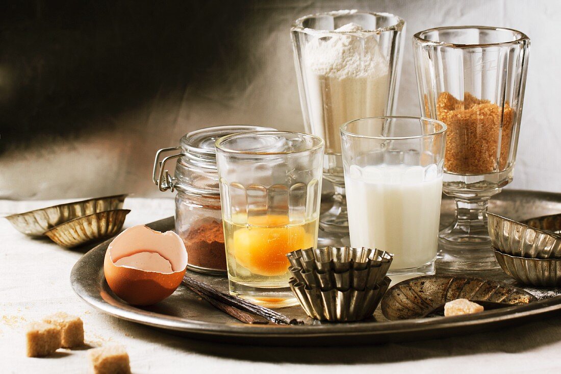 Various baking ingredients in glasses (flour, egg, brown sugar, milk) and old-fashioned cake tins on tray