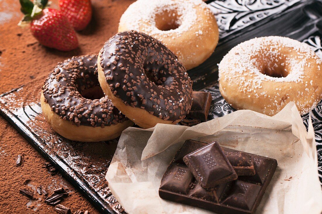 Doughnuts mit Schokoglasur und Zucker mit dunkler Schokolade auf Tablett, dahinter Erdbeeren