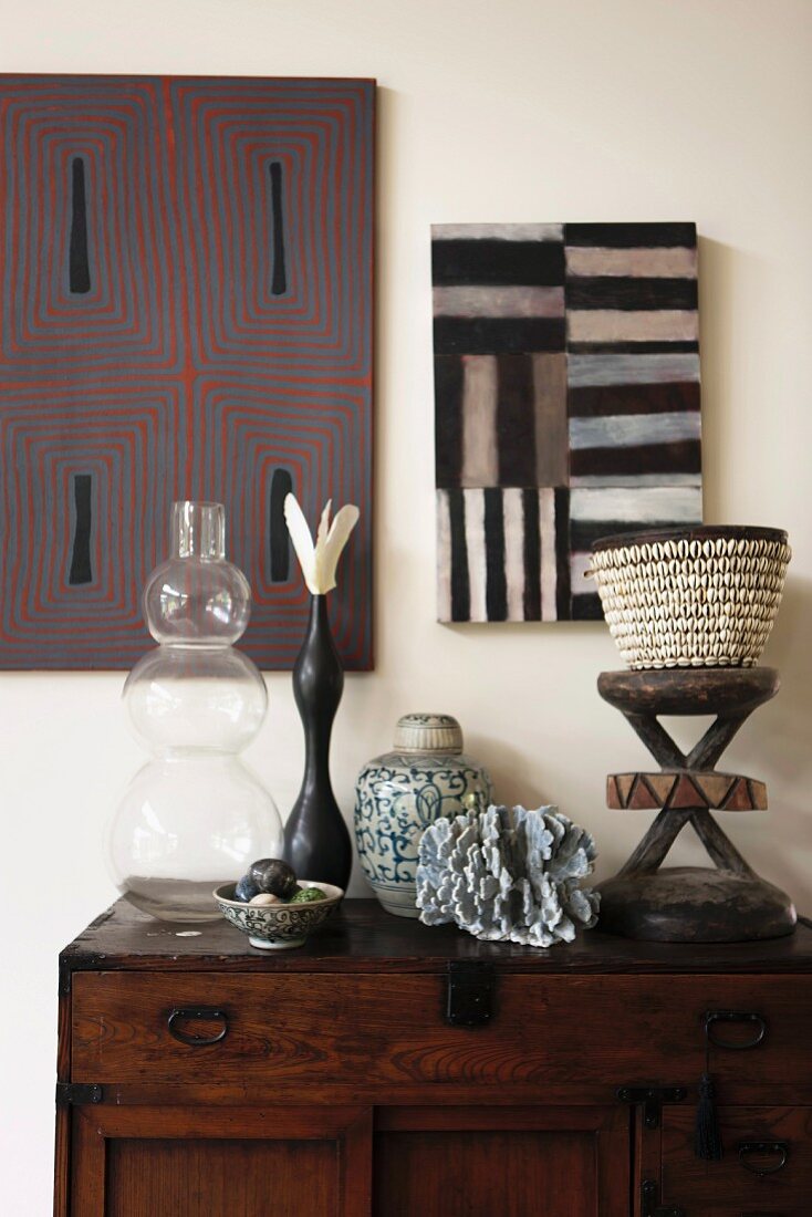 Glass vase and various artistic, African accessories on antique chest of drawers below graphic artworks on wall