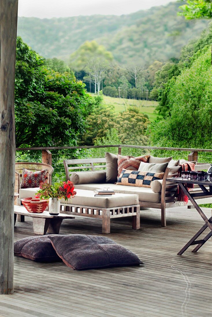 Loungeflair mit Bodenkissen auf Holzterrasse; Polstertisch und Sitzbank mit Polstern und Kissen im Hintergrund sommerliche bewaldete Hügellandschaft