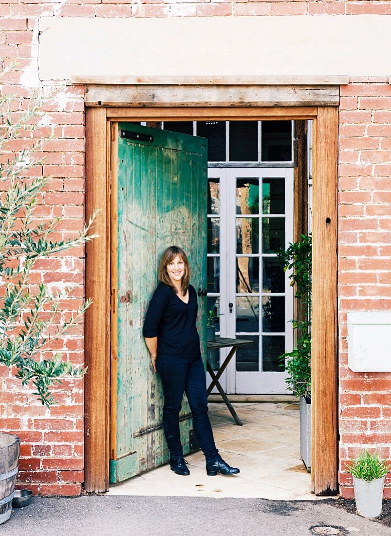 Smiling woman standing in tall, vintage front door of renovated industrial building leading into porch with white lattice door